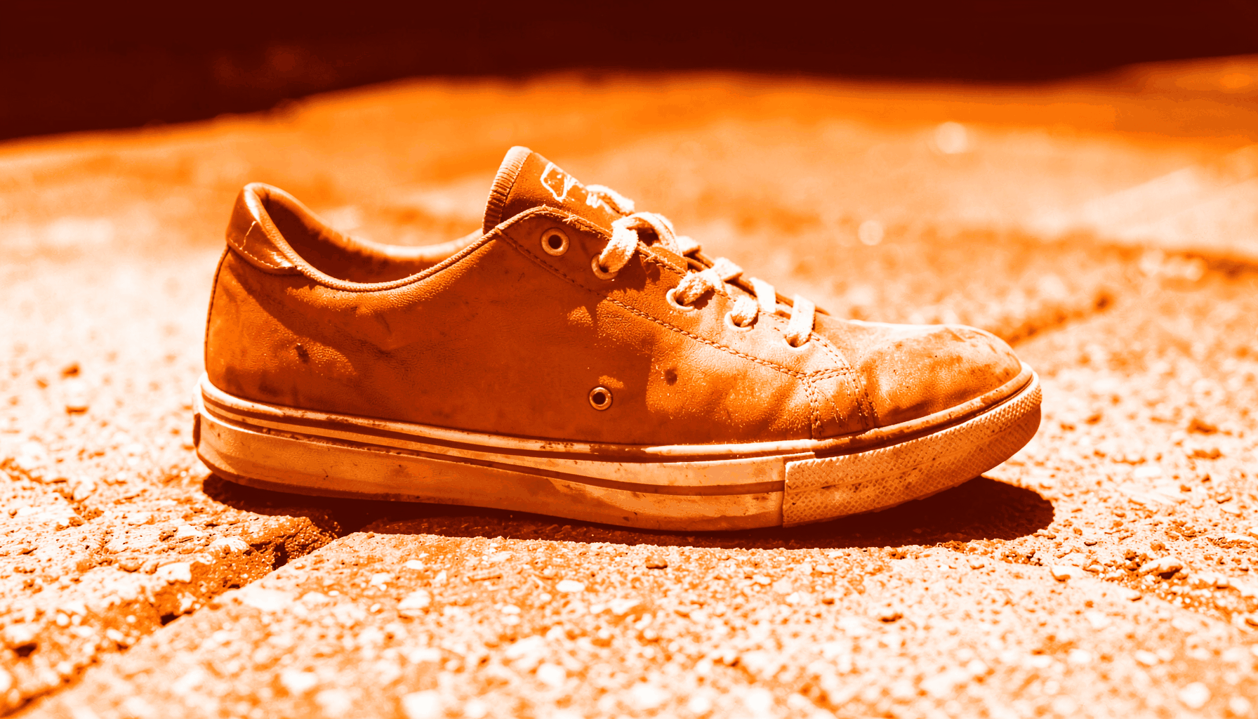 A bold duotone photograph of a single worn sneaker on sunlit pavement, rendered in burnt orange and off-white