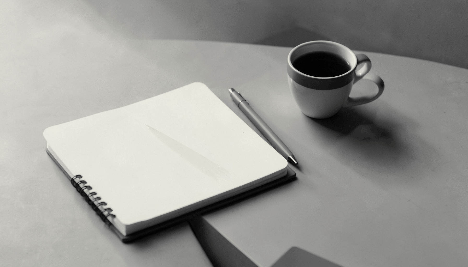 A stark duotone photograph of a minimalist desk with a notebook, pen, and morning coffee, rendered in charcoal gray and soft cream