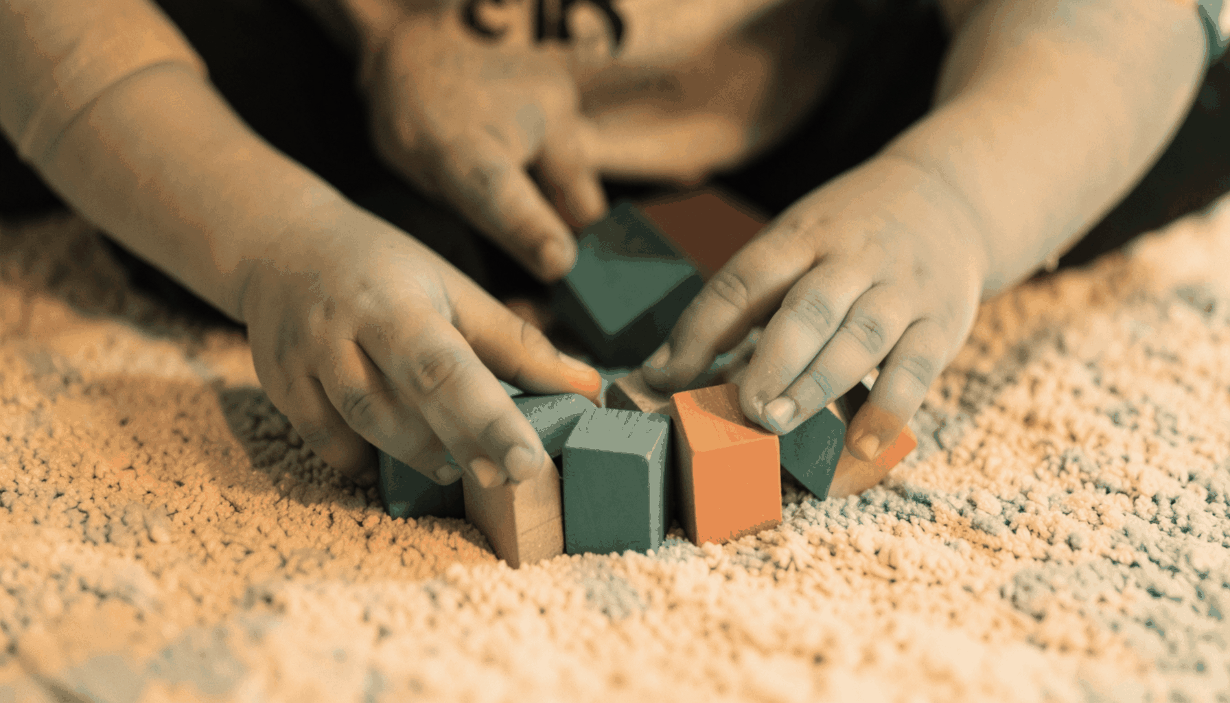 A gentle duotone photograph of a child's hands playing with colorful wooden blocks on a soft rug, rendered in warm sand and muted teal