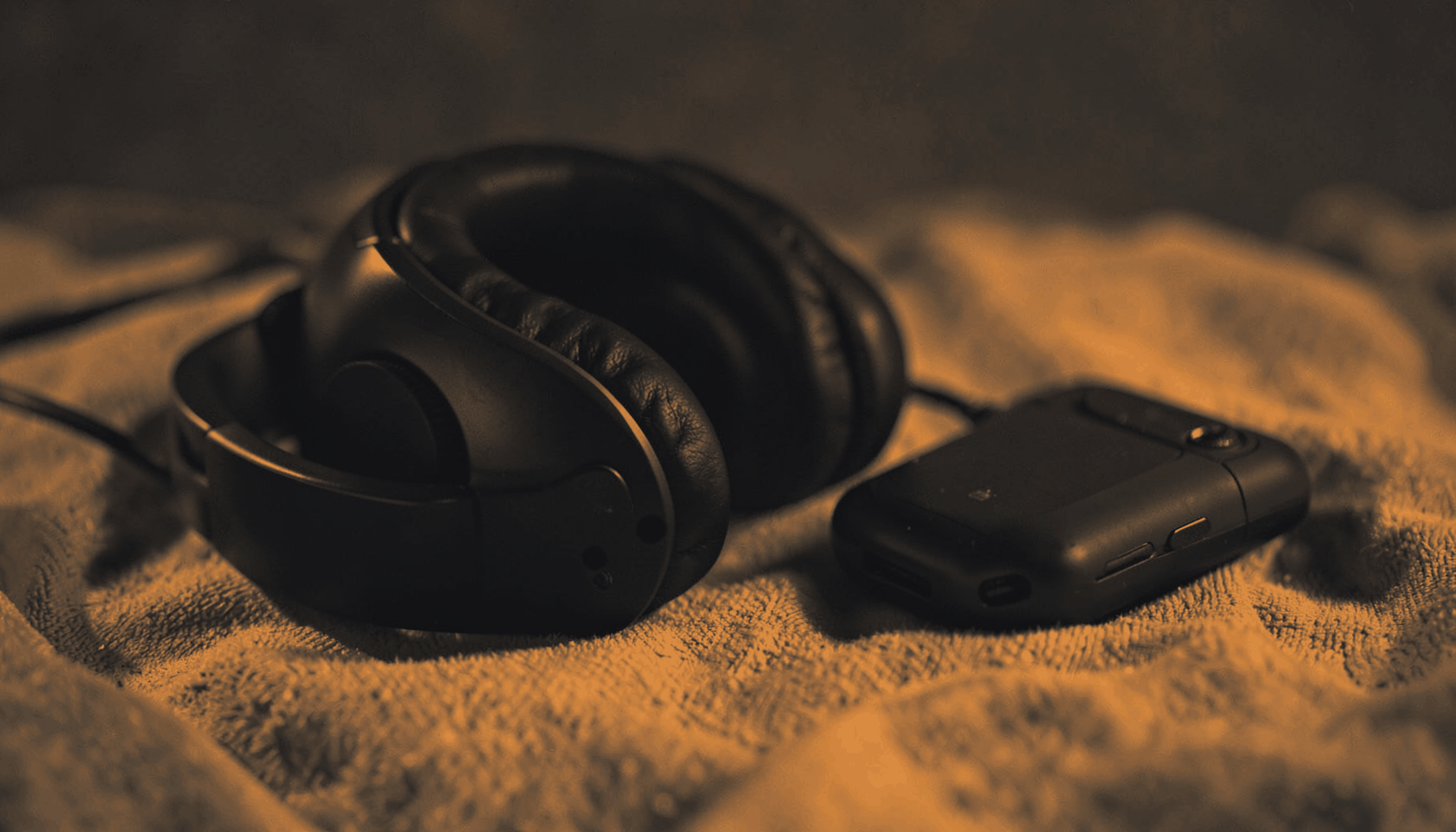 A stack of audio headphones and a portable listening device resting on a soft cloth, rendered in a hard two-tone duotone of deep charcoal and warm amber, evoking a calm, focused listening mood