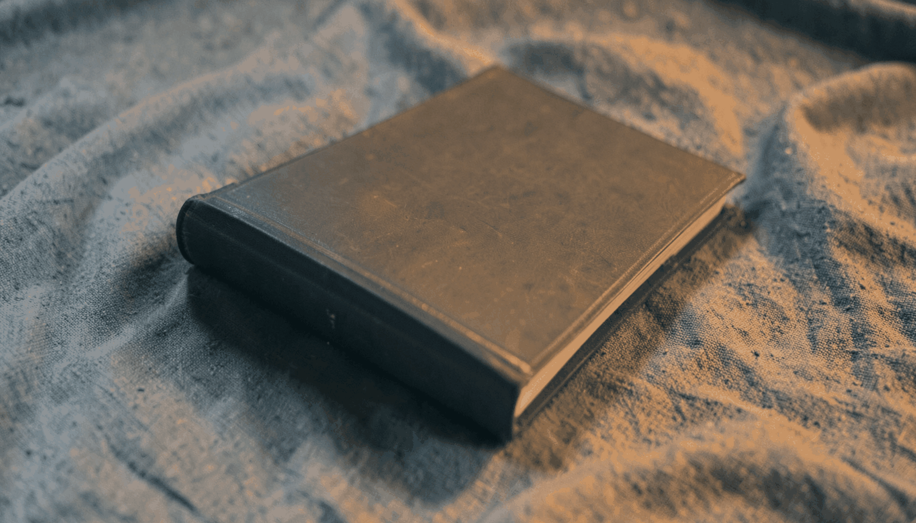 A warm duotone photograph of a novel resting on a textured fabric surface, in muted slate-blue and amber tones.