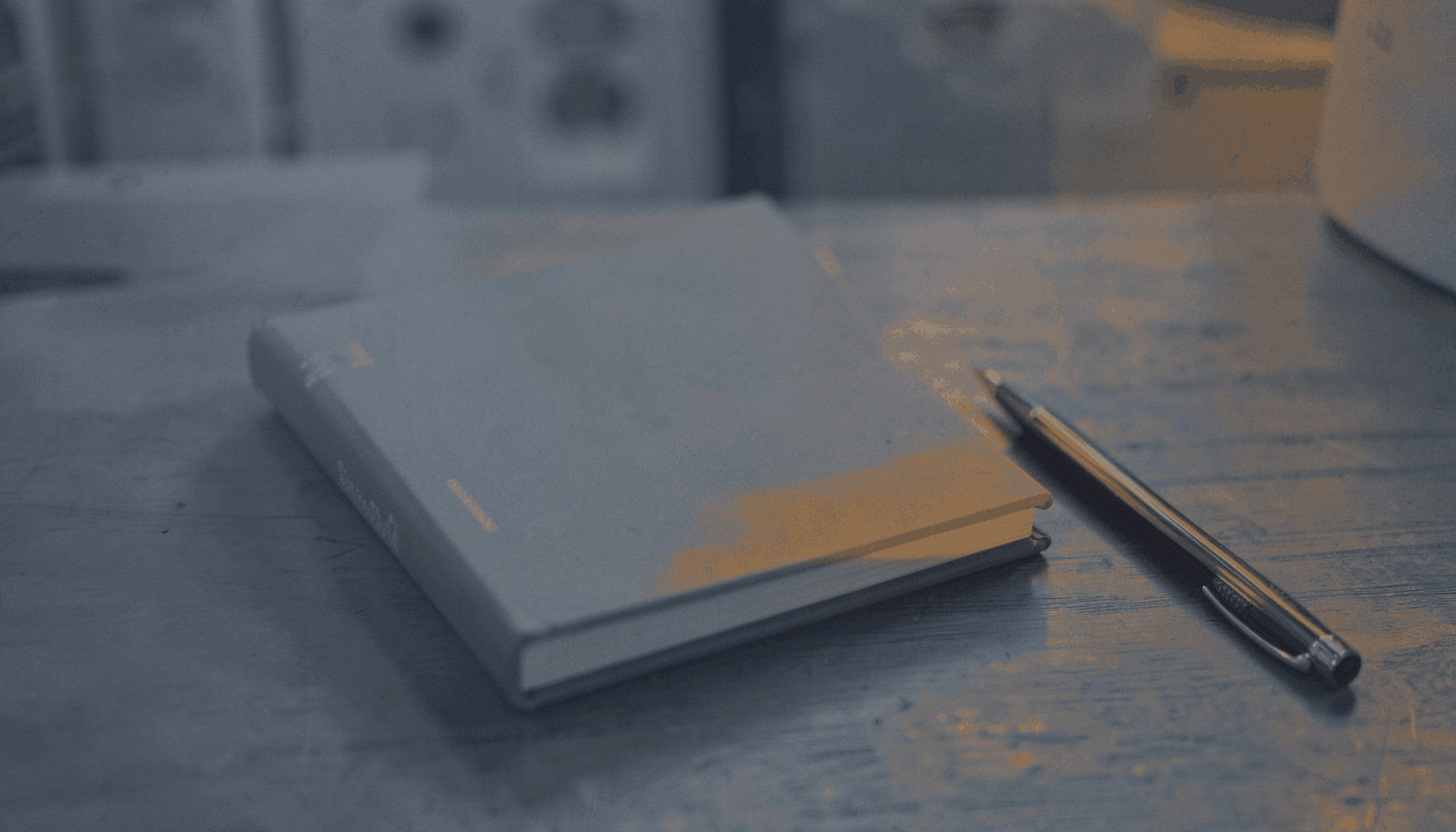 A warm duotone photograph of a business book with a pen beside it on a desk, in muted slate-blue and amber tones.